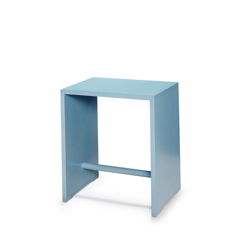 The WB Form Ulmer Stool is a minimalist blue wooden piece inspired by Max Bills design, featuring a rectangular top, open sides, and a horizontal support bar at the base. Its clean lines and simplicity stand out beautifully against a white background.