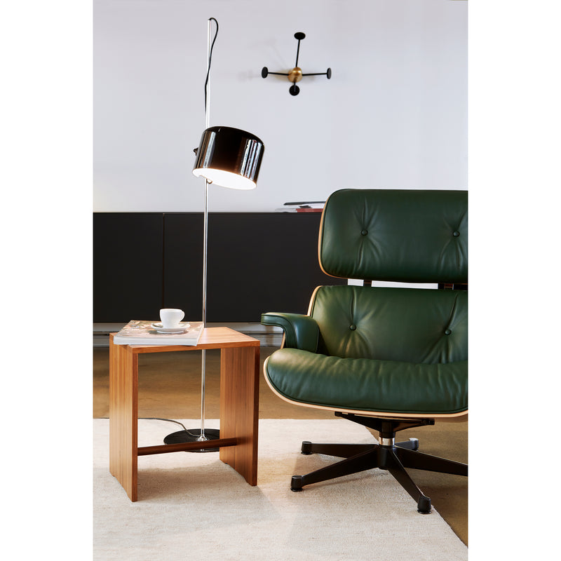 A stylish interior showcases a green leather armchair with a wooden frame, complemented by the WB Form Ulmer Stool in its nine finishes. A modern wooden side table holds a saucer and cup under a sleek floor lamps light. The backdrop includes a dark cabinet and minimalistic wall light.
