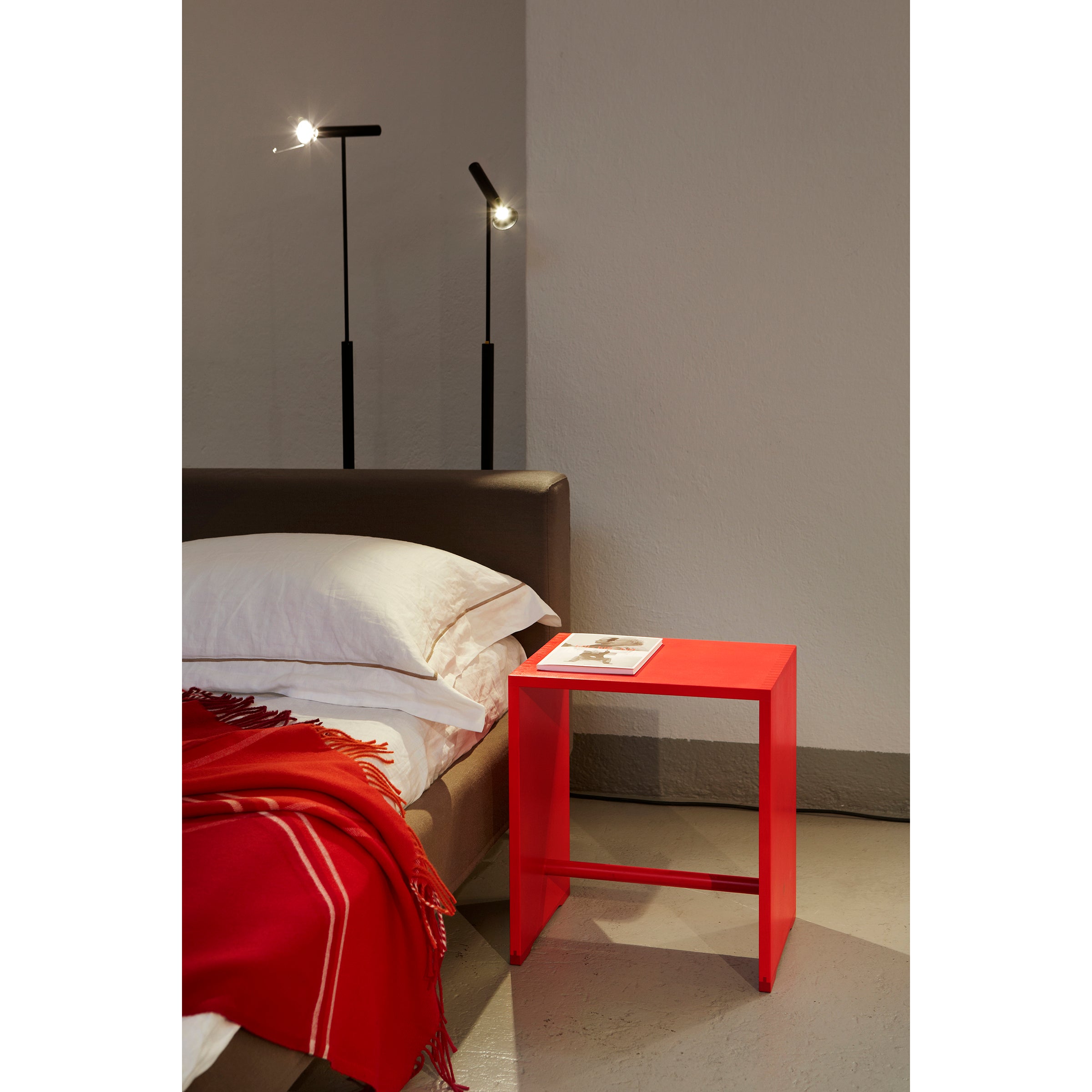 A modern bedroom features a brown bed with white pillows and a red throw blanket. An Ulmer Stool by WB Form serves as a versatile side table for magazines. In the corner, two tall black floor lamps by Max Bill illuminate the space.