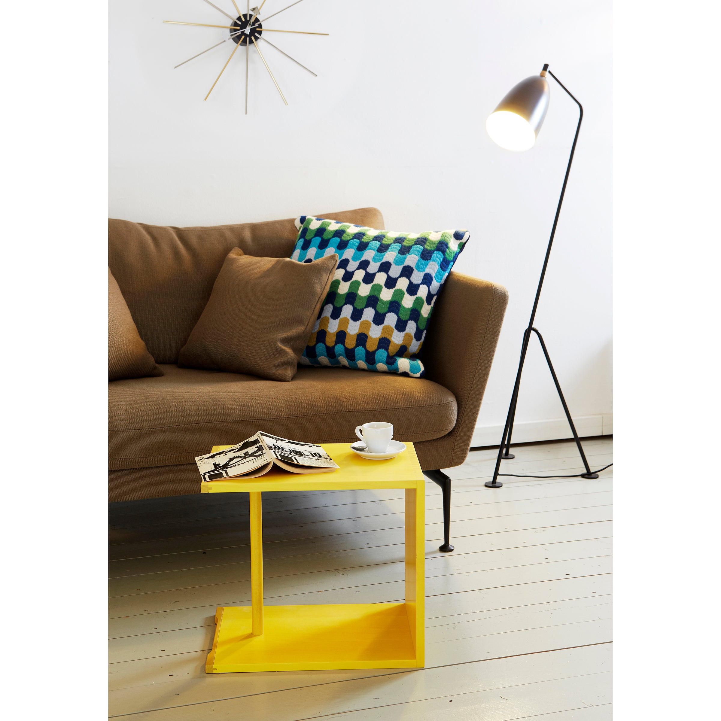 A cozy living room showcases a brown sofa, multicolored geometric cushion, and modern standing lamp. In front, a bright yellow WB Form Ulmer Stool serves as a versatile side table holding a newspaper and white cup, while a decorative clock adorns the white wall.