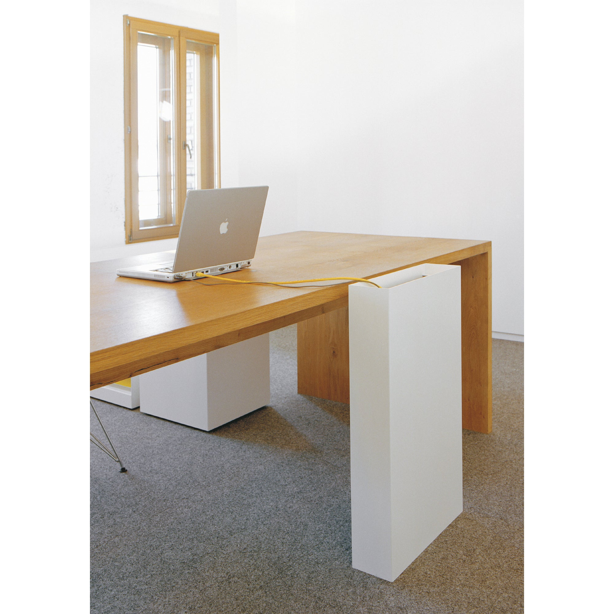 In a minimalist office, an E15 freestanding light wood table complements a laptop. Its unique design includes the BUSY wire manager, seamlessly integrating a yellow cable through a white panel at one end. A window with wooden trim completes the serene setting.
