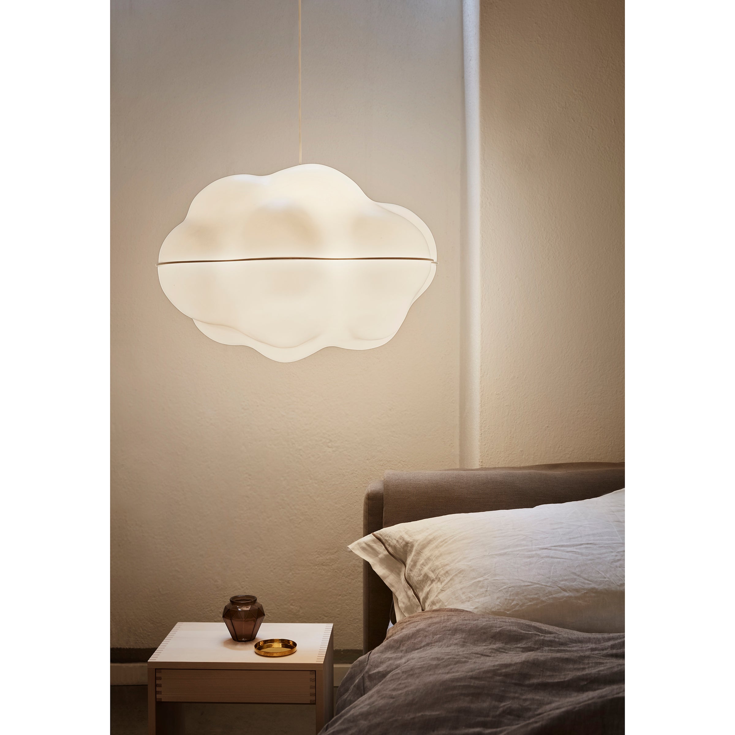 A cozy bedroom showcases WB Forms ethereal Cloud Lamp, casting a soft glow over a bedside table with a glass, book, and gold dish. A gray headboard and white bedding create a serene contrast against the beige wall.