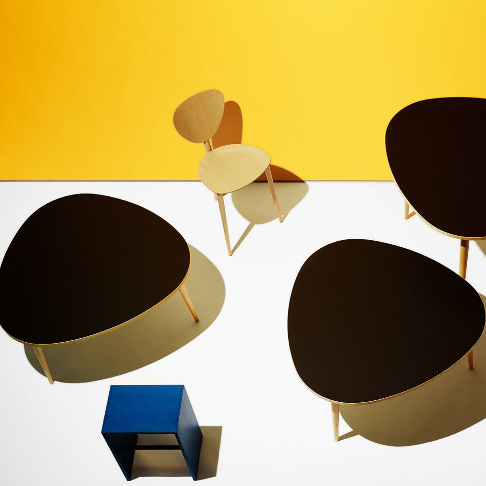 A vibrant room with a bold yellow wall showcases modern furniture, featuring three black tables with light wood legs around a minimalist WB Form Dreibein Chair. A small blue cube rests on a white floor, casting clear shadows.