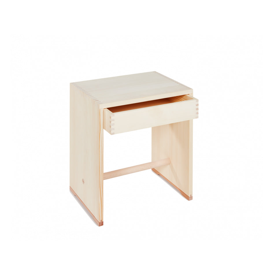 The WB Form Ulmer Stool with Drawer is a minimalist wooden side table featuring an open drawer, dovetail joints, and a natural finish. Its functional elegance, highlighted by a simple base and horizontal support beam, is reminiscent of designs by Hans Gugelot and Max Bill.