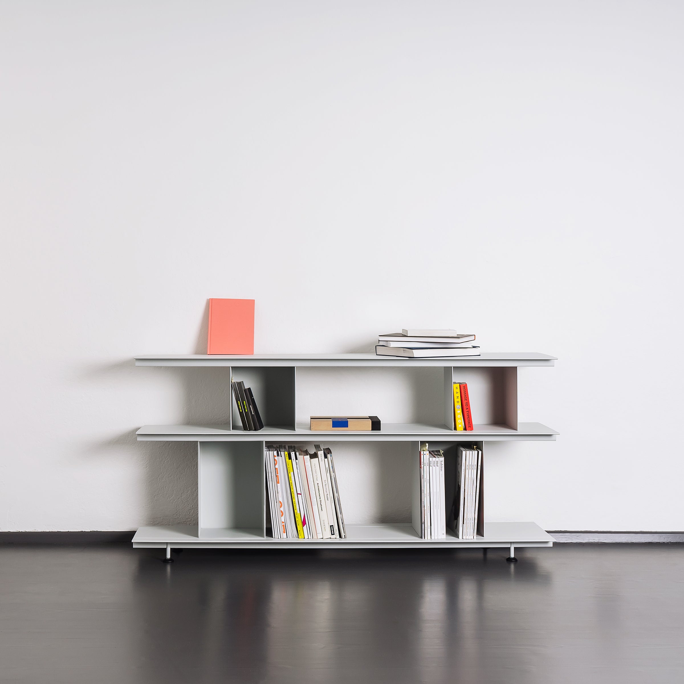 The Zoll D by Moormann, part of a modular system, is a minimalist bookshelf with three shelves against a white wall. It holds books and magazines, some stacked and some upright. The light-colored furniture contrasts beautifully with the dark floor, highlighting its versatile design.