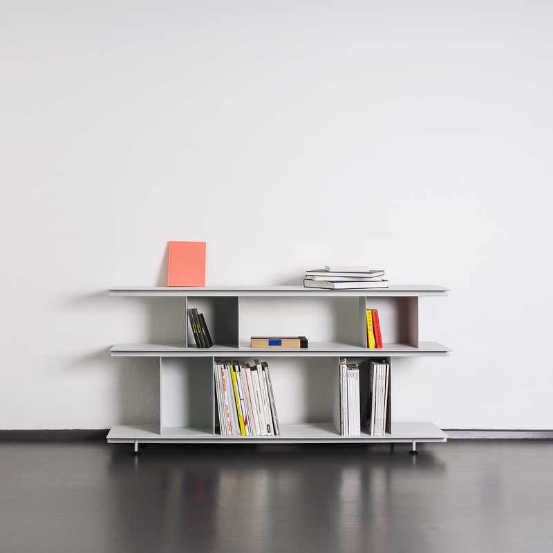 The Zoll D by Moormann, part of a modular system, is a minimalist bookshelf with three shelves against a white wall. It holds books and magazines, some stacked and some upright. The light-colored furniture contrasts beautifully with the dark floor, highlighting its versatile design.