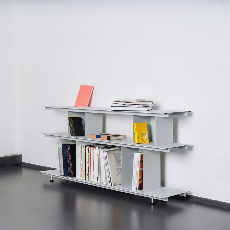 The Moormann Zoll D, a minimalist white bookshelf, features three shelves against a plain white wall. This versatile piece neatly displays books and colored notebooks, with open spaces for an organized look. Its tool-free assembly design complements the dark gray floor seamlessly.