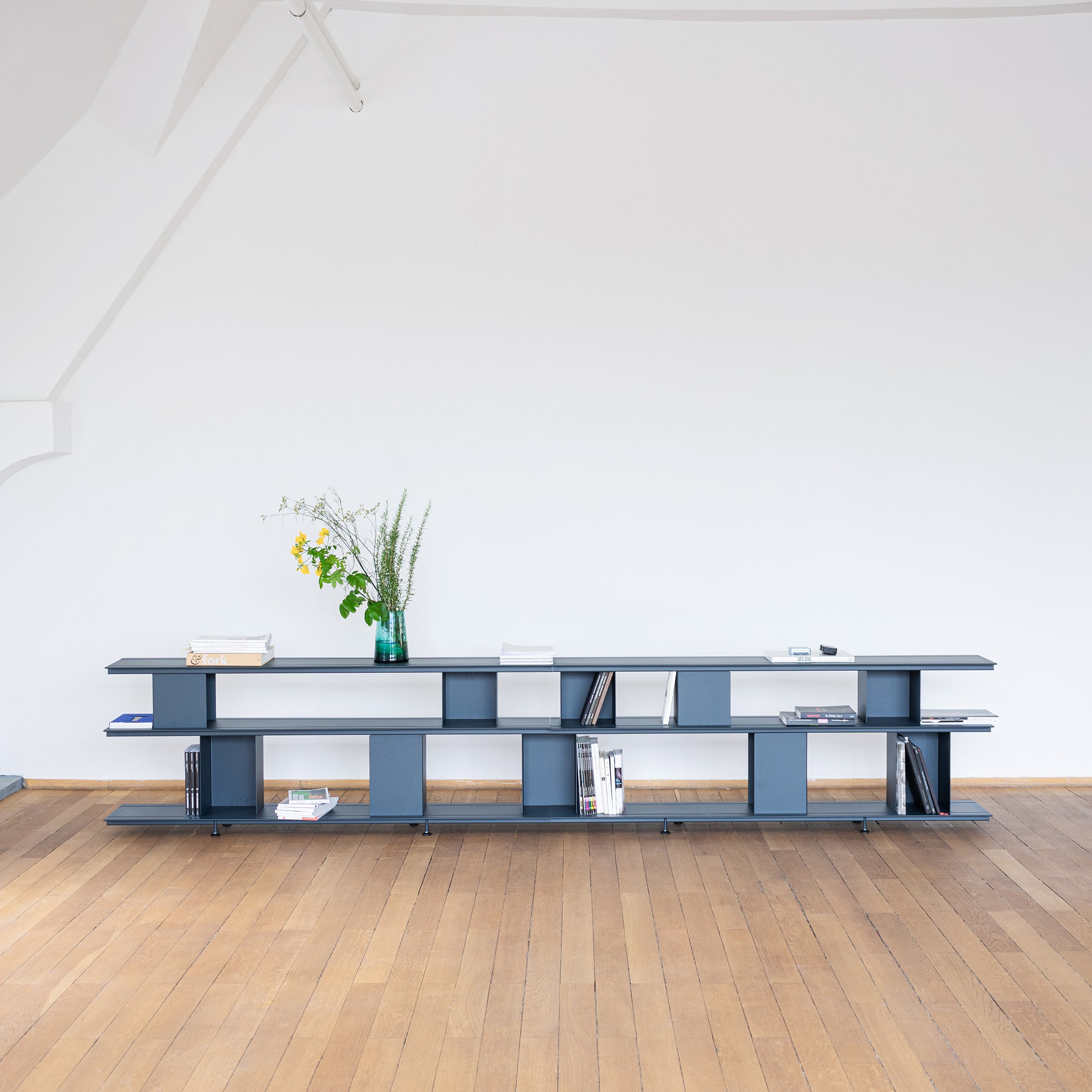 The Zoll D by Moormann is a modern, long, open-shelf unit displaying books and a green vase with flowers against a white wall. This versatile furniture piece offers tool-free assembly. Its wooden floor and minimalist design create a clean, spacious look.