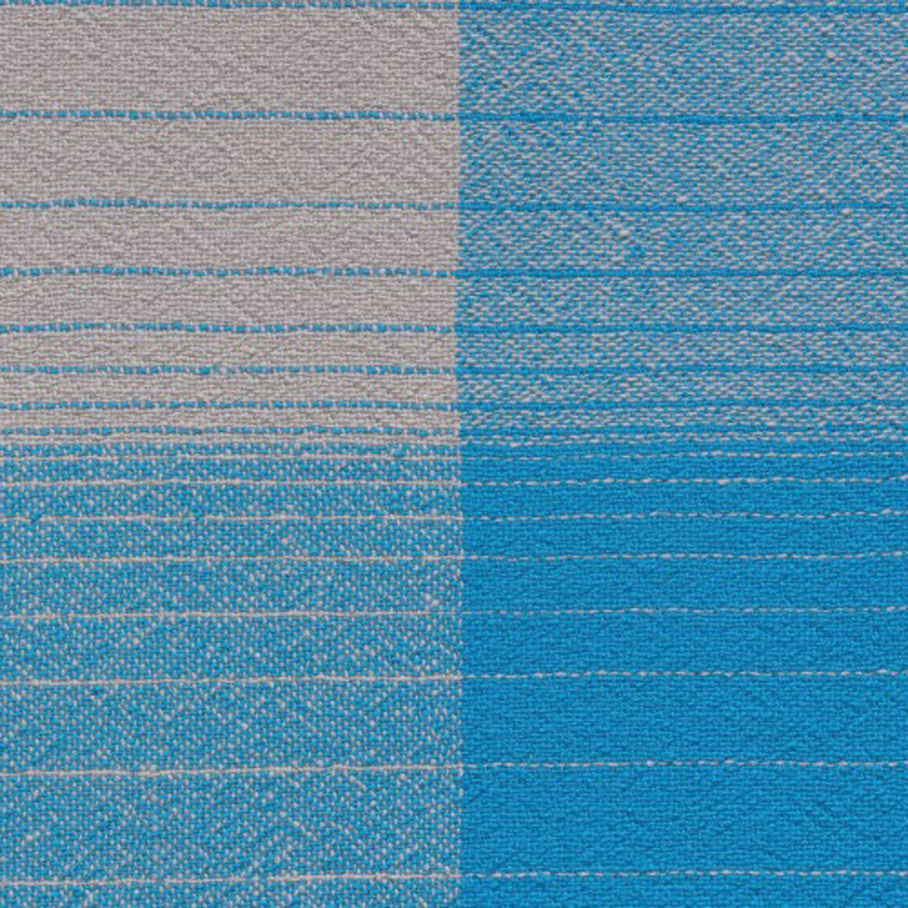 A close-up of the AC13 CUADRO blanket by E15 shows its hand-woven design: the left section is light grey with slim blue stripes, while the right is vibrant blue with fine horizontal lines, showcasing a refined texture and color contrast in merino wool.