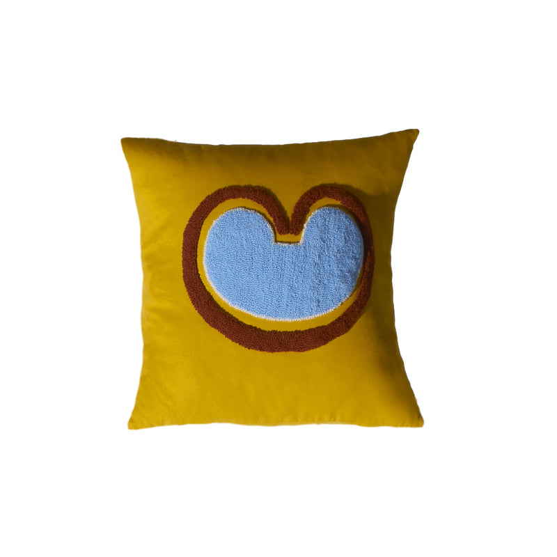 The Ames Nido Coraz by Ames is a square yellow pillow inspired by Sebastian Herkner’s Nido cushions, featuring a raised textured center with a large blue shape bordered in brown and red.