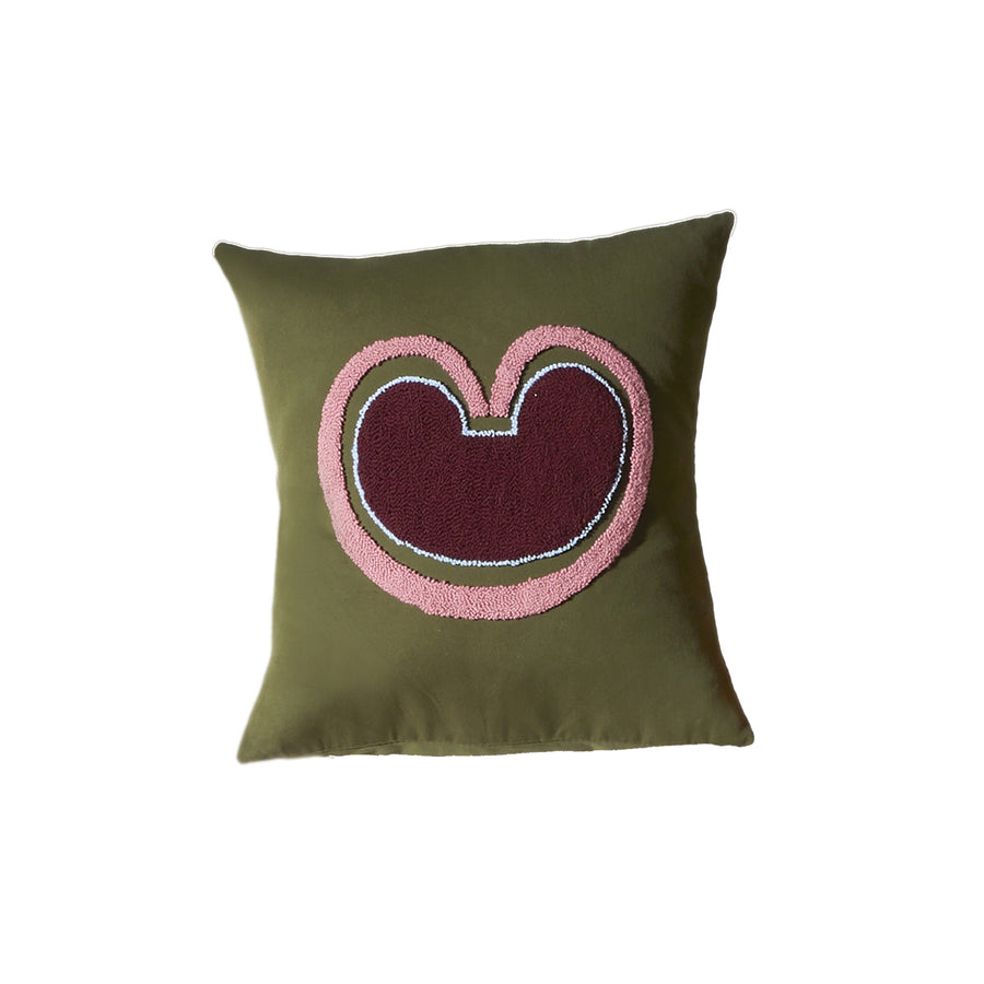 The Ames Nido Coraz pillow by Ames is a square olive green cushion from the Nido collection, featuring an abstract central design with maroon, pink, and white shapes resembling a stylized bean or heart outline.