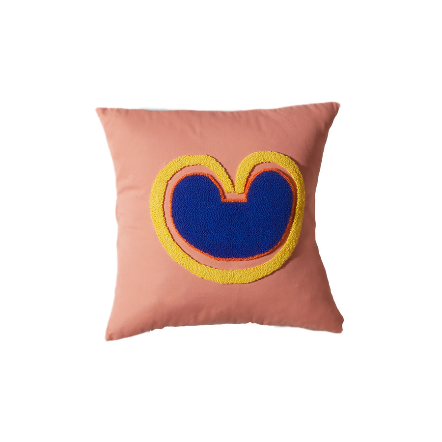 The Ames Nido Coraz cushion is a square pink pillow featuring a handmade abstract design with a blue shape outlined in yellow and orange, resembling a stylized heart or rounded M.