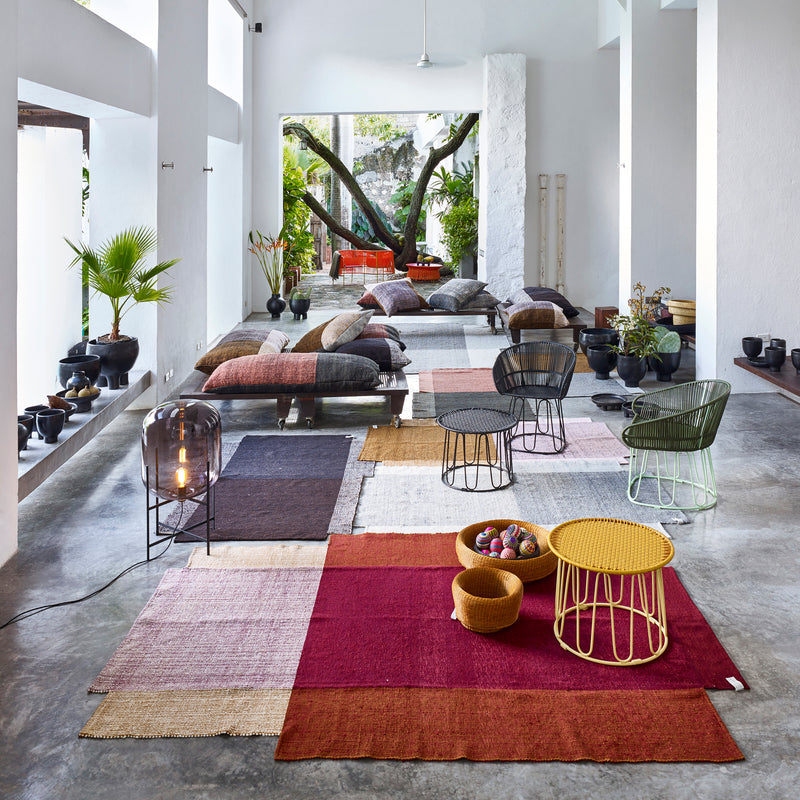 In a contemporary, light-filled living space, Ames colorful Nobsa Rugs and cushions brighten a polished concrete floor. Wire-frame chairs encircle a small round table, with potted plants enhancing the decor. Expansive windows offer views of lush greenery outside.