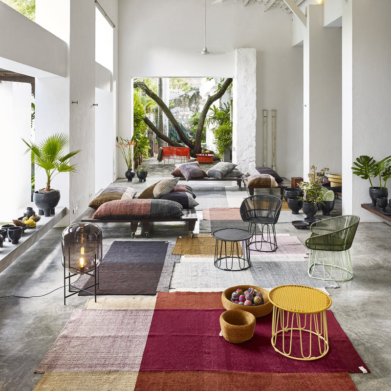 A bright, modern living space is furnished with the Ames Circo Side Table by Ames, designed by Sebastian Herkner. Colorful rugs, low sofas piled with cushions, and lush potted plants create a relaxed room filled with natural light.