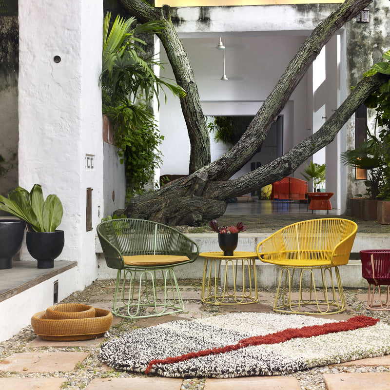 Outdoor patio with colorful woven chairs from the CIRCO collection, Ames Circo Side Tables by Ames, a textured rug, potted plants, and a large tree beside a white building, creating a modern and inviting setting.