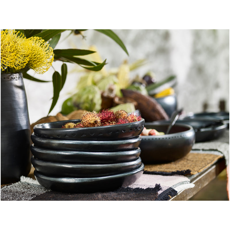 Ames Barro Plate, a dark ceramic beauty, sits on a woven mat displaying its unique craftsmanship. Nested within are rambutan fruits. Behind them, a black vase with yellow flowers and green leaves enhances the scene alongside other bowls and dishes.