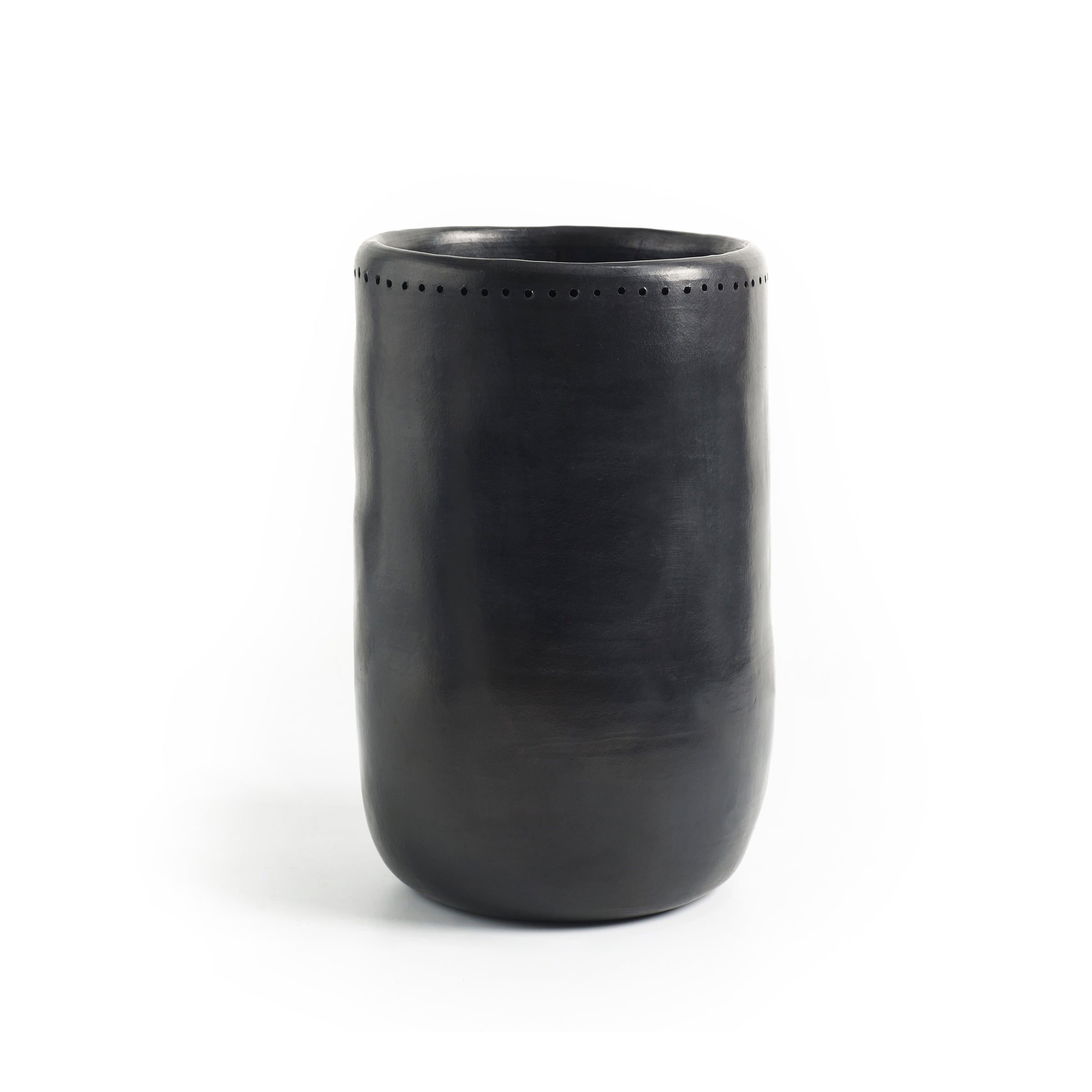 Set against a plain white background, the Barro Vase 1 by Ames is a tall, smooth, black ceramic vase with a slightly tapered shape and small decorative perforations near the top. Exhibiting exquisite Colombian craftsmanship, it reflects the artistry of handmade pottery.
