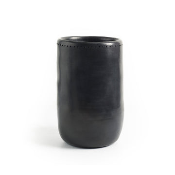Set against a plain white background, the Barro Vase 1 by Ames is a tall, smooth, black ceramic vase with a slightly tapered shape and small decorative perforations near the top. Exhibiting exquisite Colombian craftsmanship, it reflects the artistry of handmade pottery.