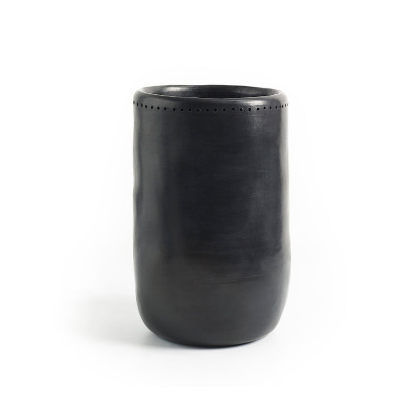 Set against a plain white background, the Barro Vase 1 by Ames is a tall, smooth, black ceramic vase with a slightly tapered shape and small decorative perforations near the top. Exhibiting exquisite Colombian craftsmanship, it reflects the artistry of handmade pottery.