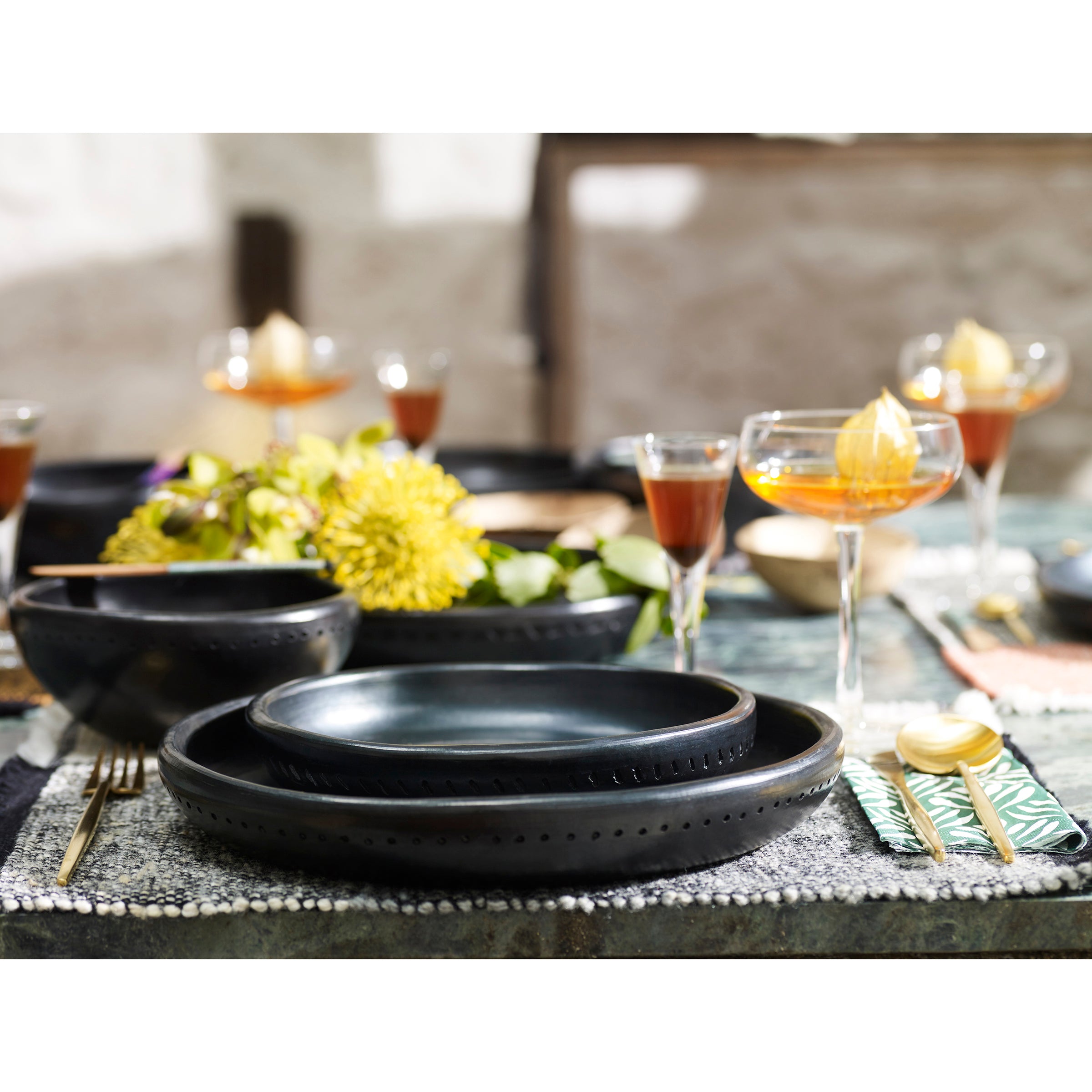 Elegant dinner setting featuring Ames Barro ceramic plates, gold utensils, and glasses of amber liquid. A centerpiece with yellow flowers in traditional pottery and greenery accents the table, covered by a textured placemat.