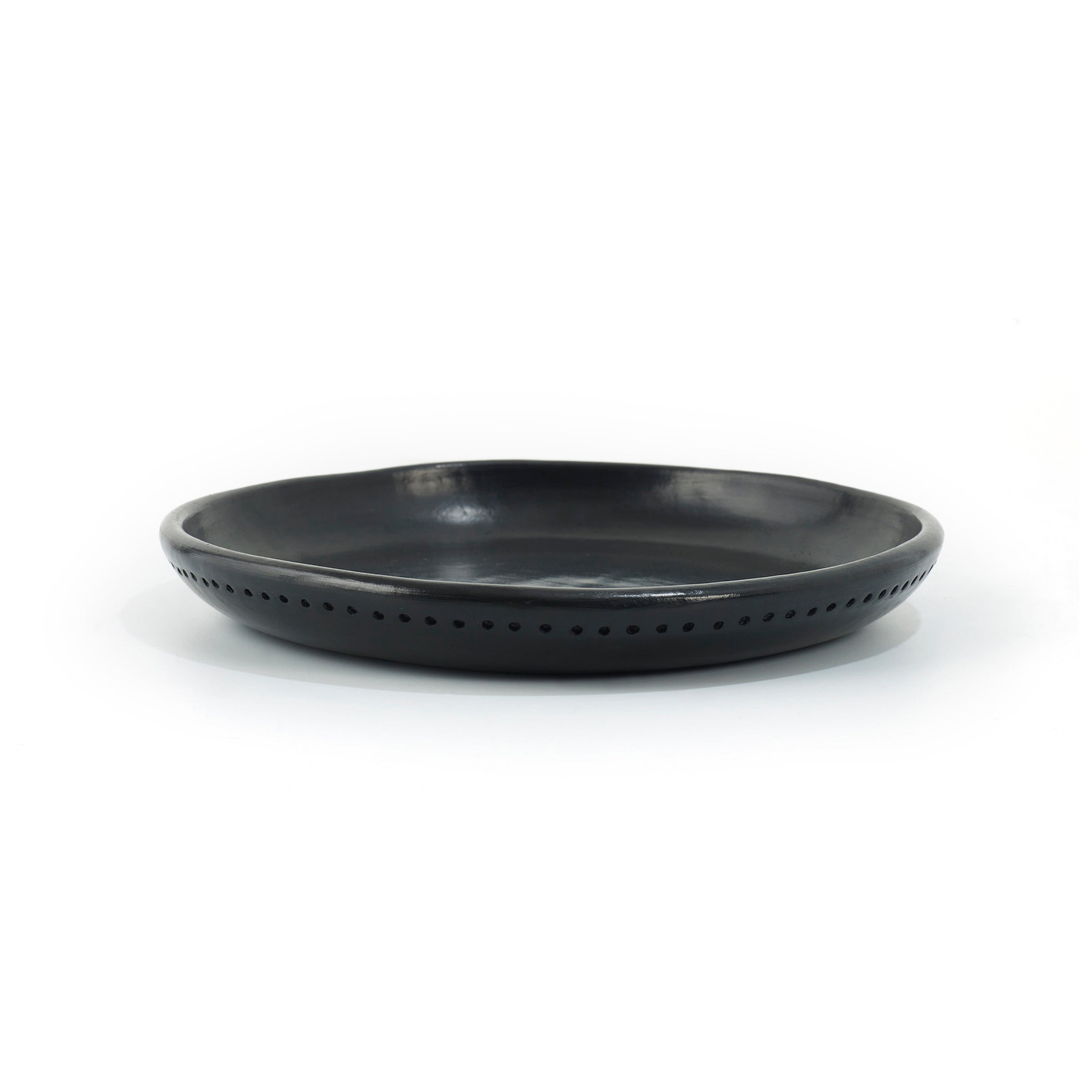 The Barro Plate by Ames is a black, round ceramic dish with a slightly raised rim and tiny perforations along its edge, evoking traditional pottery against a plain white background.