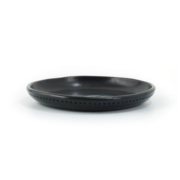 The Barro Plate by Ames is a black, round ceramic dish with a slightly raised rim and tiny perforations along its edge, evoking traditional pottery against a plain white background.