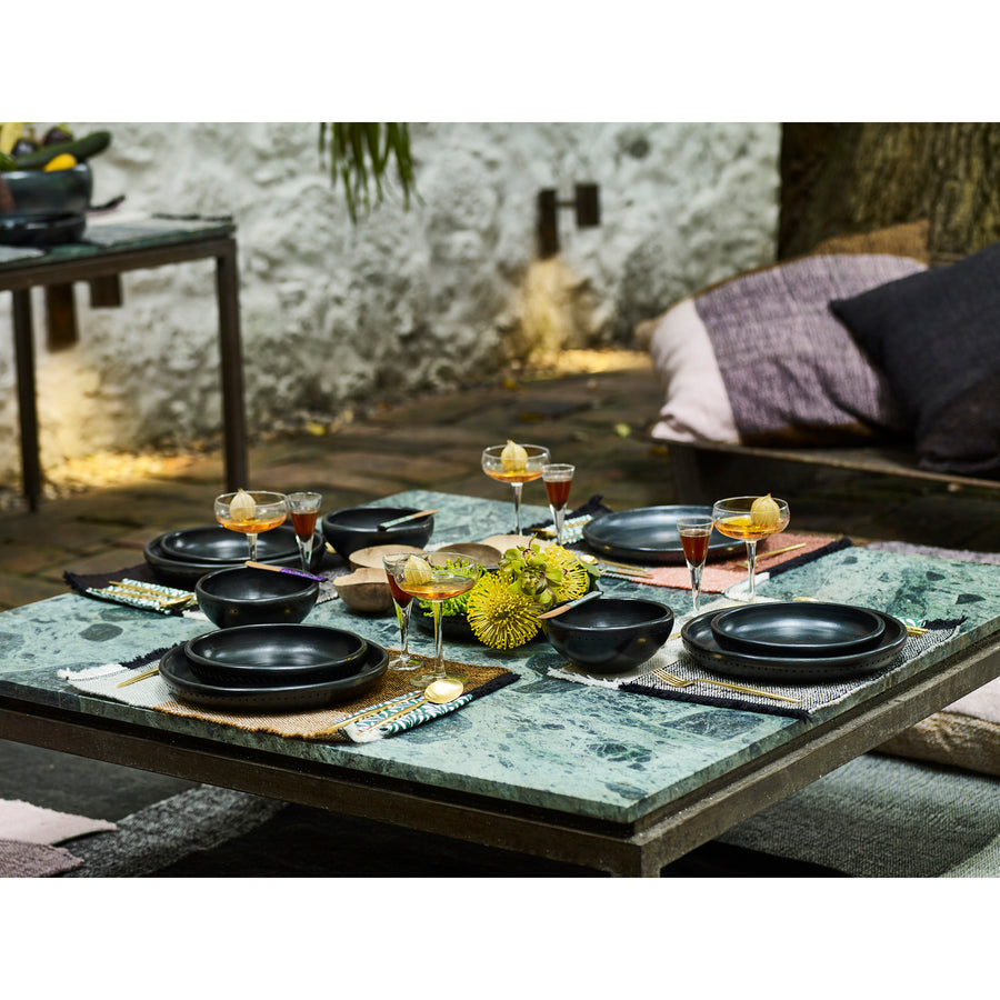 Amess Barro Plates grace a stone outdoor table set with unique ceramics, including black dishes and bowls. Yellow flowers steal the spotlight as the centerpiece, flanked by cocktails in glasses. Cushions offer seating while plant containers and lush foliage create a vibrant background against the stone wall.