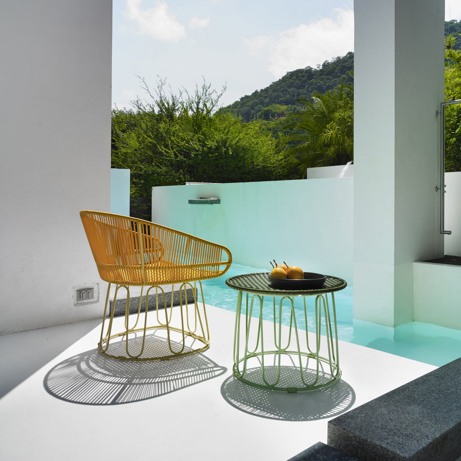 A yellow chair and the Ames Circo Side Table by Ames, topped with two drinks, sit on a white patio beside a turquoise pool, surrounded by greenery and mountains beneath a partly cloudy sky.