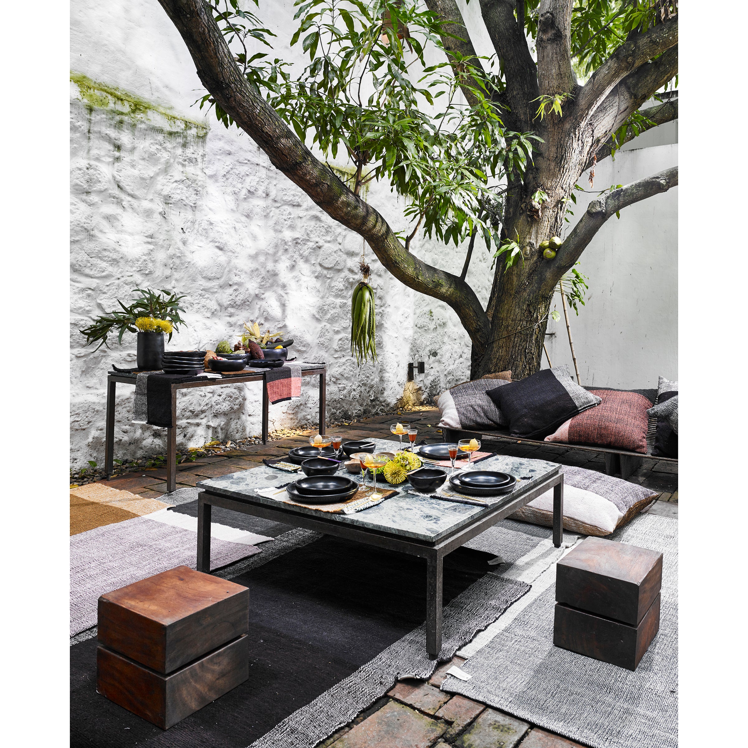 A cozy outdoor dining area under a leafy tree features a low table set with black dinnerware, cushions including Ames Nobsa Cushions by Ames for seating, a side table with plants, and textured rugs on stone flooring against a white stone wall.