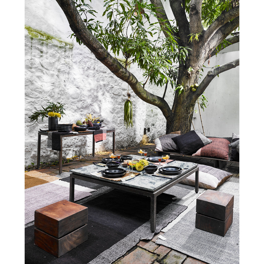 A cozy outdoor dining area under a leafy tree features a low table set with black dinnerware, cushions including Ames Nobsa Cushions by Ames for seating, a side table with plants, and textured rugs on stone flooring against a white stone wall.