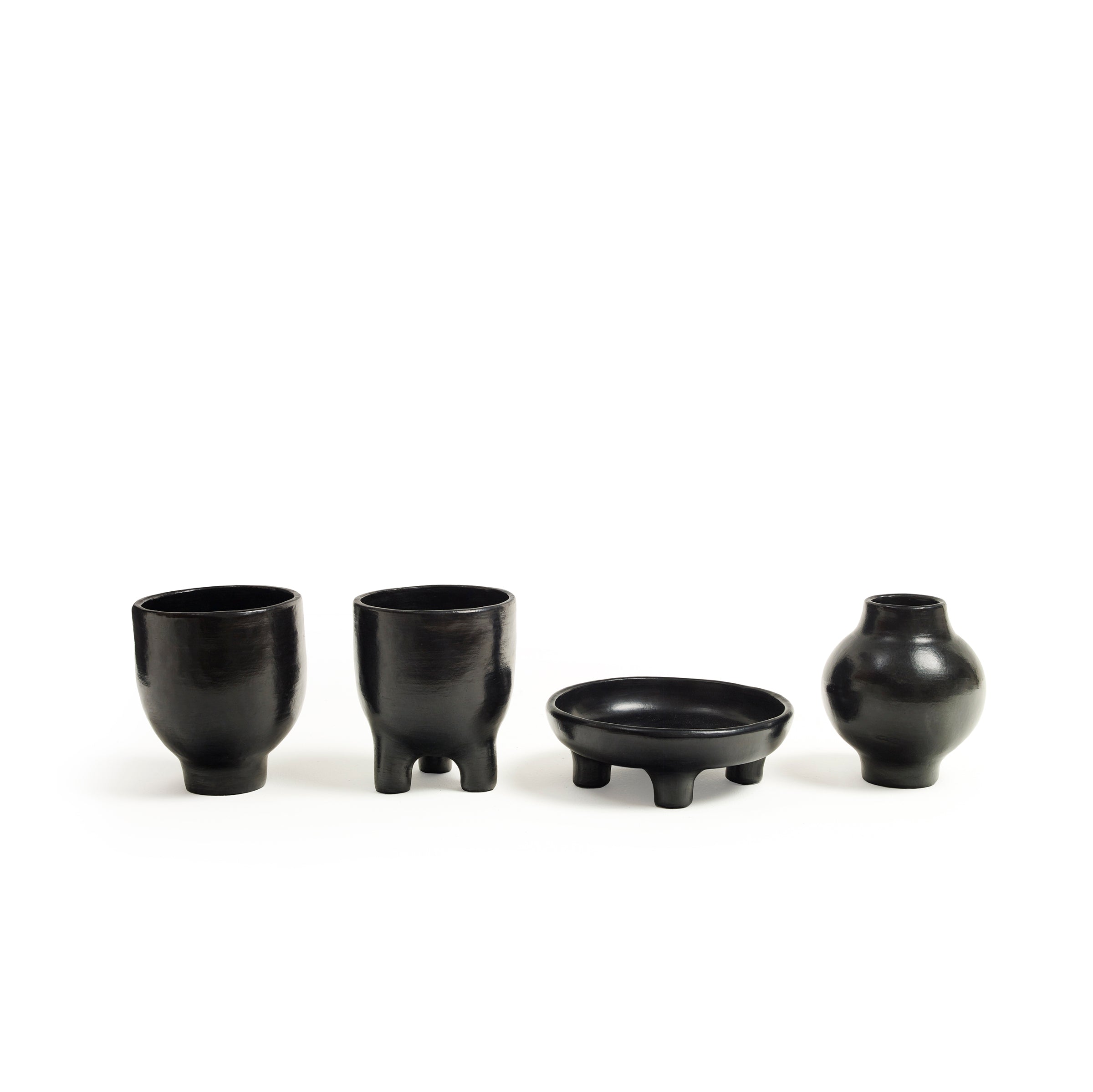 Explore the beauty of Colombian pottery with Ames: two black Barro Pot 2 cups on short legs, a three-legged shallow dish, and a small rounded vase.