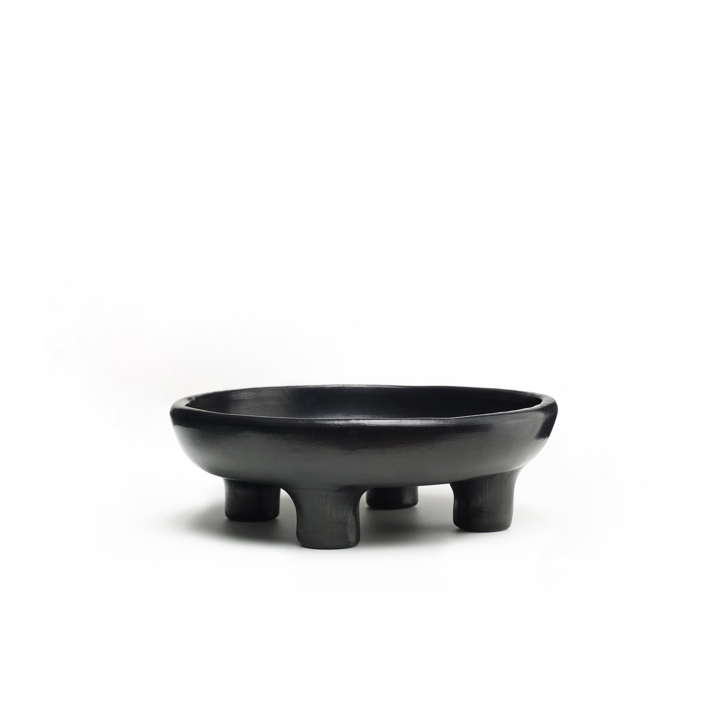 The Ames Barro Tray 1 by Ames is a black, round, shallow ceramic bowl with four short, sturdy legs—an elegant piece of handmade Colombian ceramics—set against a plain white background.