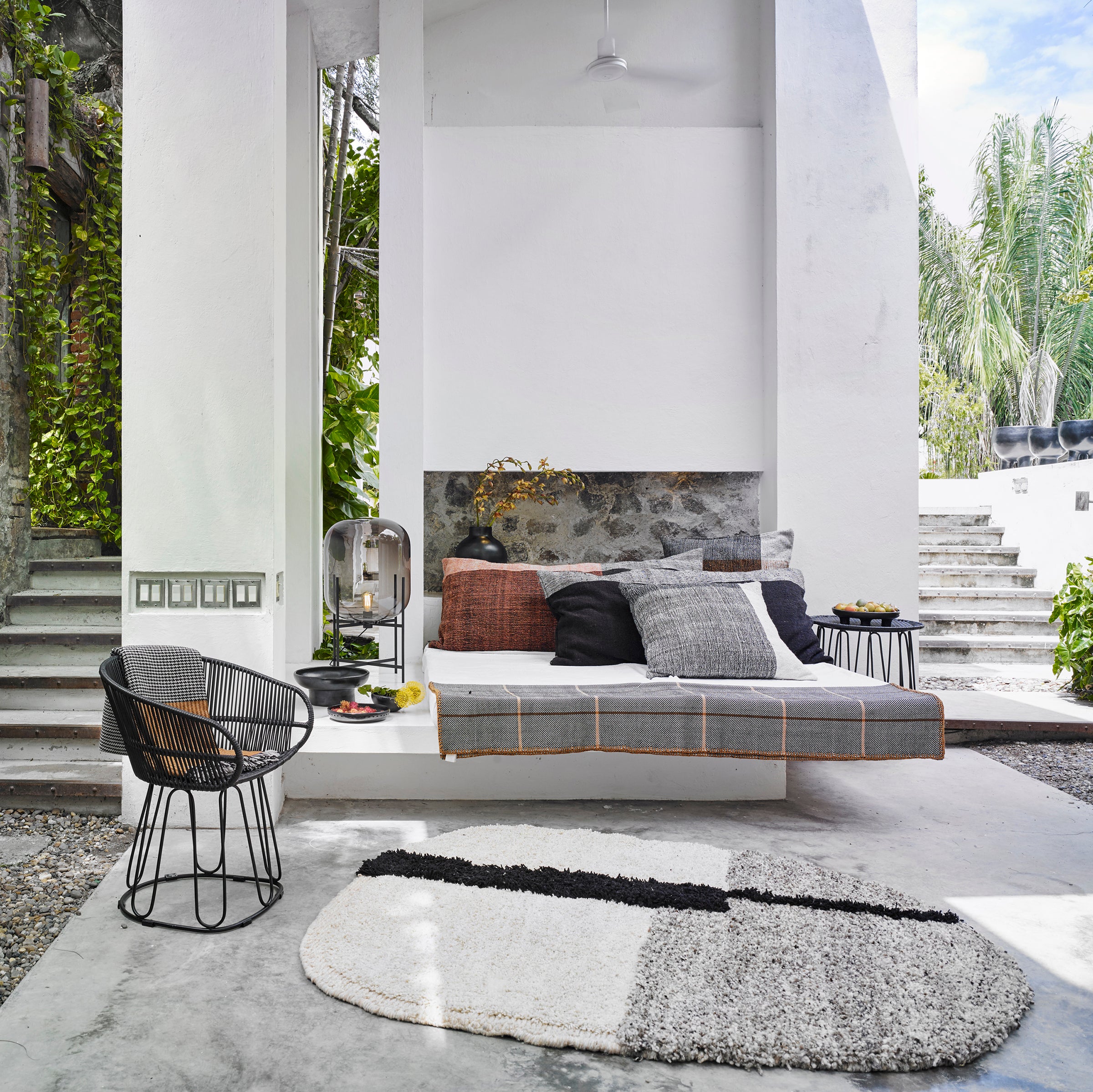 Modern outdoor seating area with a gray cushioned bench, Ames Nobsa Cushions by Ames, a small round table, wire chair, textured round rug, and lush greenery visible through the open structure. Bright and airy ambiance.