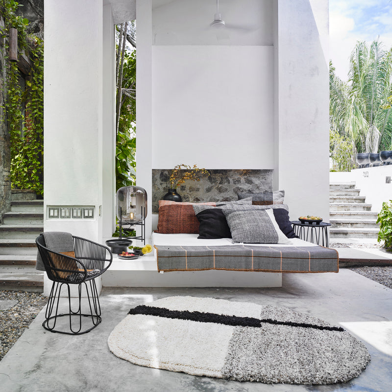 Modern outdoor seating area with a gray cushioned bench, Ames Nobsa Cushions by Ames, a small round table, wire chair, textured round rug, and lush greenery visible through the open structure. Bright and airy ambiance.