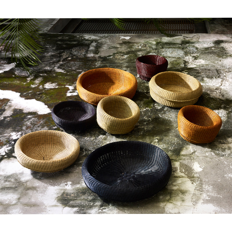 Eight Ames Fibra Baskets by Ames, handcrafted in Colombia from esparto grass in black, tan, brown, orange, and yellow earth tones and assorted sizes, arranged on mossy concrete with green foliage overhead.