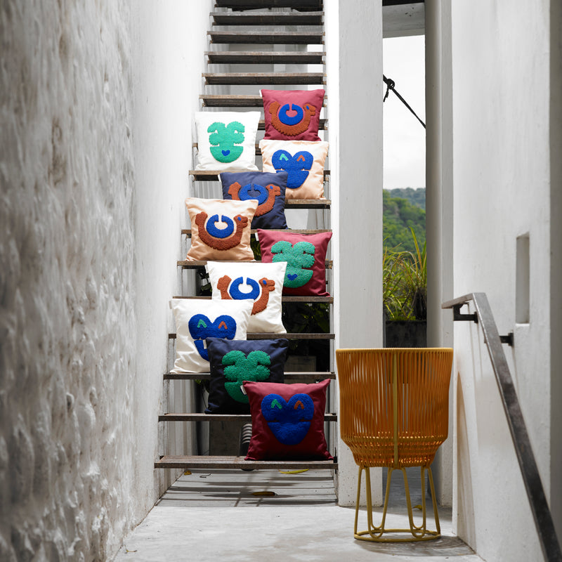 Nido Qeztal throw pillows by Ames, adorned with playful abstract face designs in green, blue, and brown, are arranged on outdoor stairwell steps. A yellow wire-frame chair is positioned at the bottom right, set against a lush greenery backdrop reminiscent of Museo del Oro.