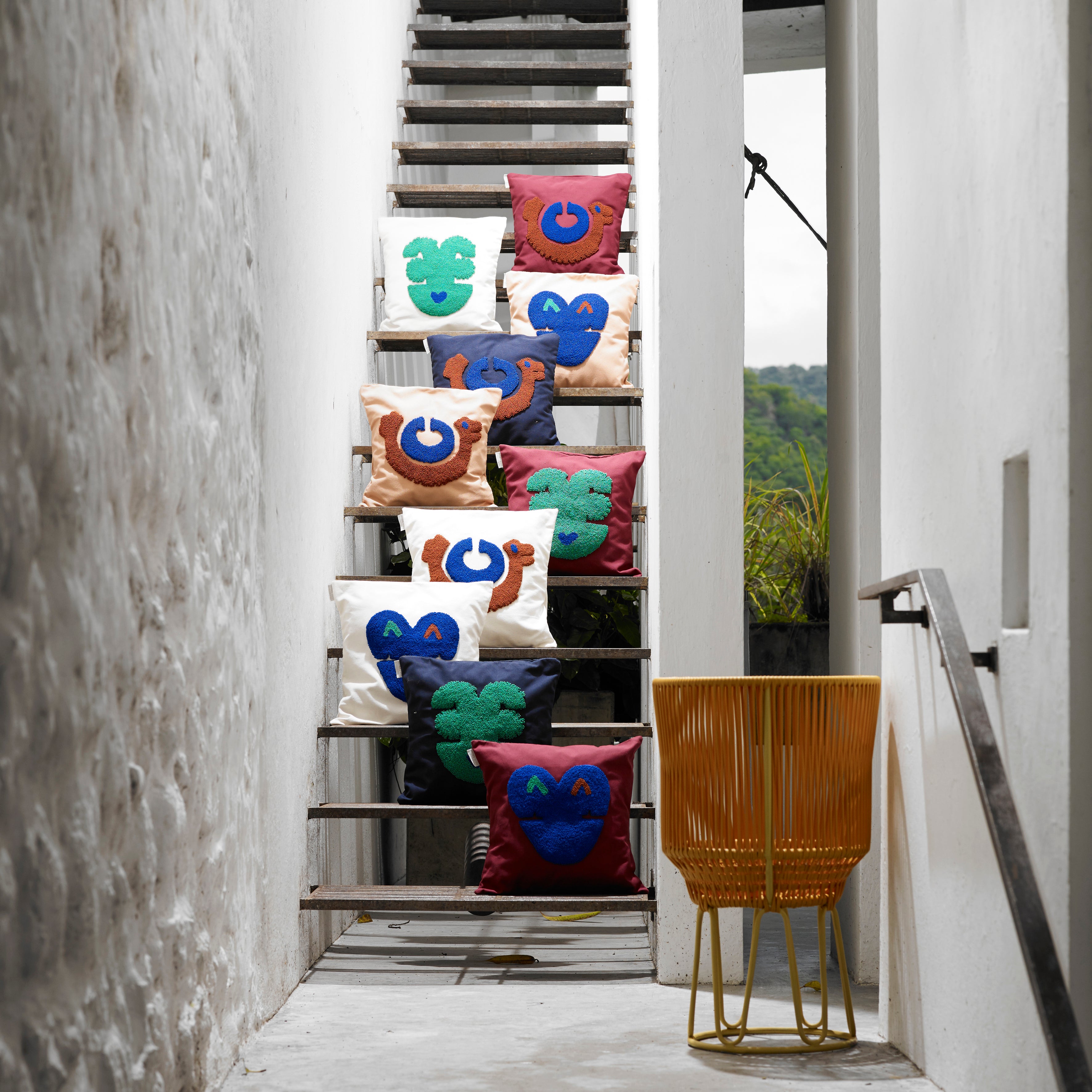 Nido Kukuy pillows by Ames, adorned with cheerful faces, are displayed on a narrow stairway framed by white walls. A yellow wire chair at the base invites relaxation. Lush greenery peeks through an upper corner opening, creating a serene backdrop for this cozy scene.