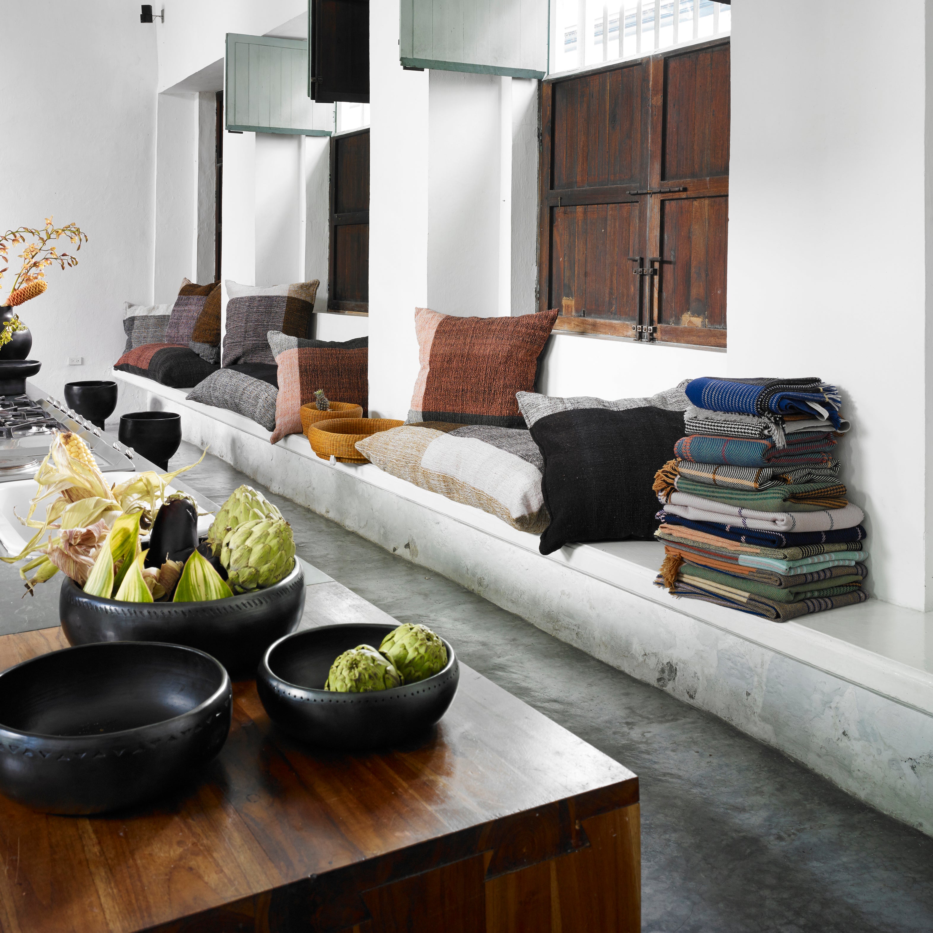 A modern, minimalist living space features a built-in bench with assorted pillows, an Ames Manta Plaid by Ames draped on one end, and a wooden table set with black bowls and artichokes in the foreground.