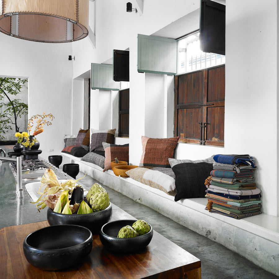 A bright, modern kitchen features a long bench under large windows, decorated with hand-woven Ames Nobsa Cushions by Ames and stacked textiles. Black bowls with artichokes rest on a wooden table, while indoor plants bring a fresh touch.
