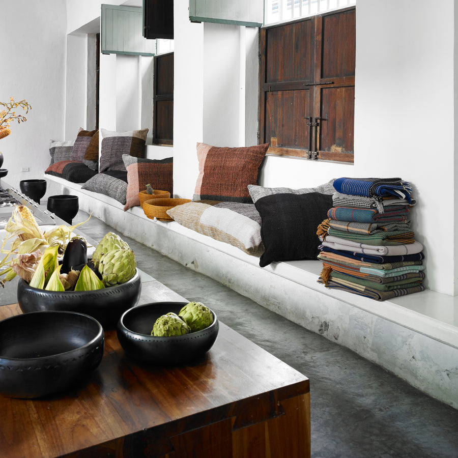 A modern, minimalist room features a long white bench with colorful pillows, stacked woolen blankets, Ames Mulera Throws by Ames, wooden windows, and a table topped with black bowls of artichokes and decor.