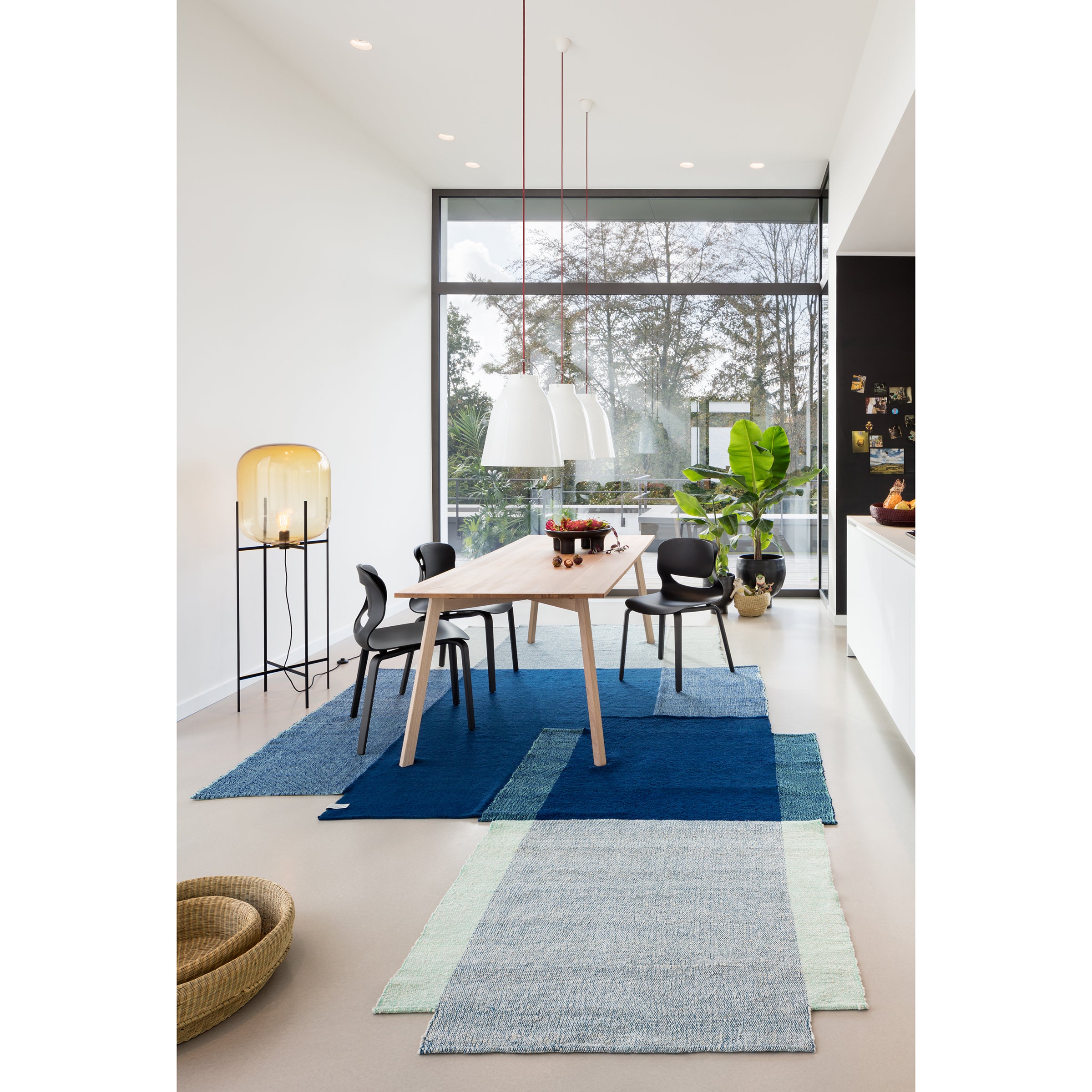 A modern dining room features a sleek wooden table, black chairs, and layered Ames Nobsa rugs in blue and gray. Pendant lights are overhead, while a large window showcases greenery outside. A stylish lamp and potted plant accentuate the space, highlighting its hand-woven wool elegance.