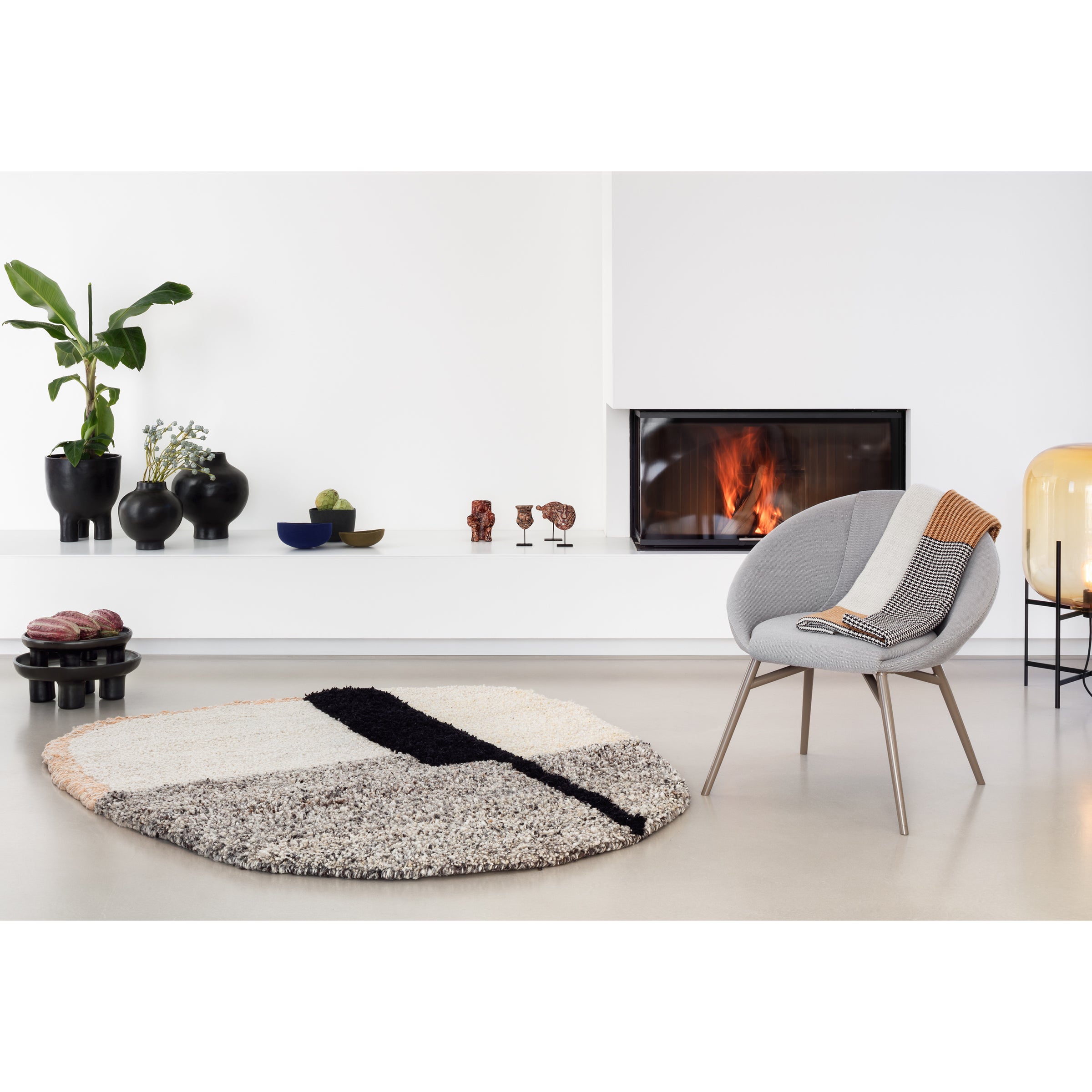 A modern living room showcases a gray chair with a patterned throw, an Ames Nudo Rug, and a lit fireplace. Black and blue vases with decorative items grace the white shelf. A plant and yellow lamp add depth, enhancing the spaces Colombian arts and crafts flair.