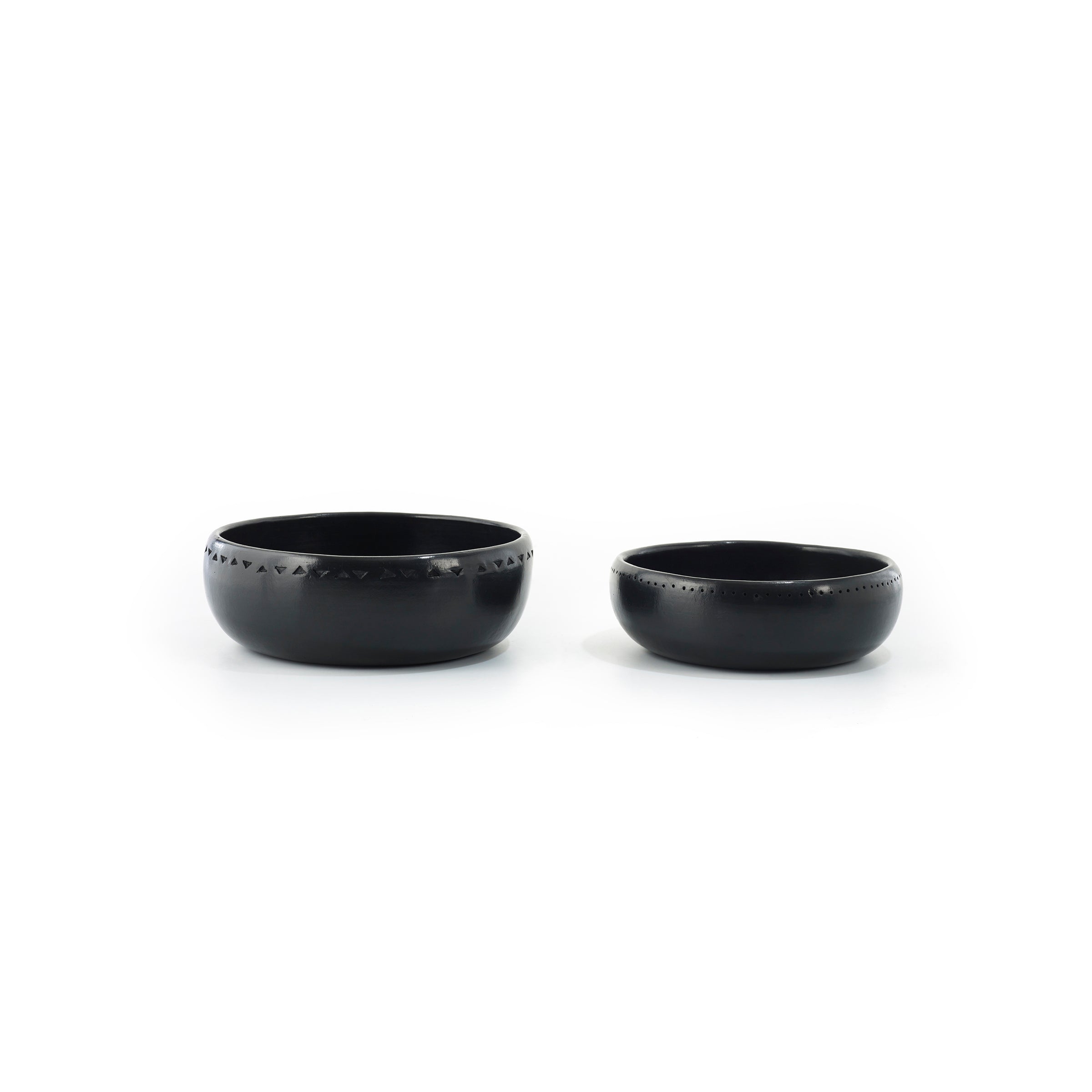 Two Ames Barro Bowls, crafted in matte black ceramic with varying sizes and etched geometric patterns around the top edge, are displayed side by side on a plain white background, highlighting traditional pottery artistry.