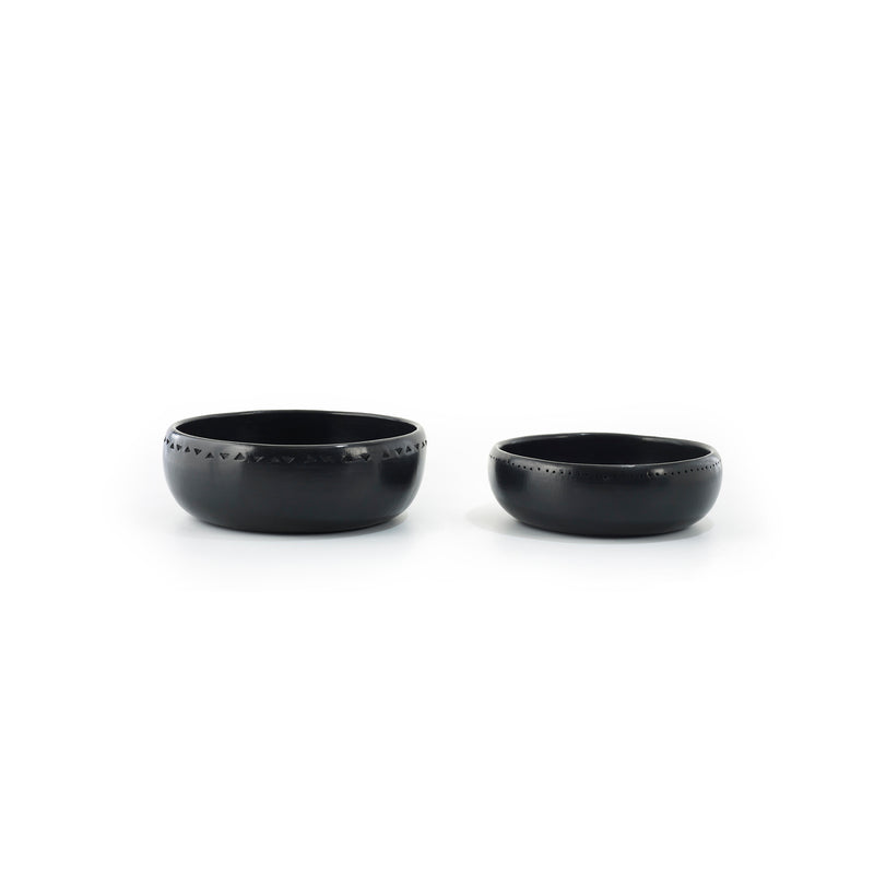 Two Ames Barro Bowls, crafted in matte black ceramic with varying sizes and etched geometric patterns around the top edge, are displayed side by side on a plain white background, highlighting traditional pottery artistry.