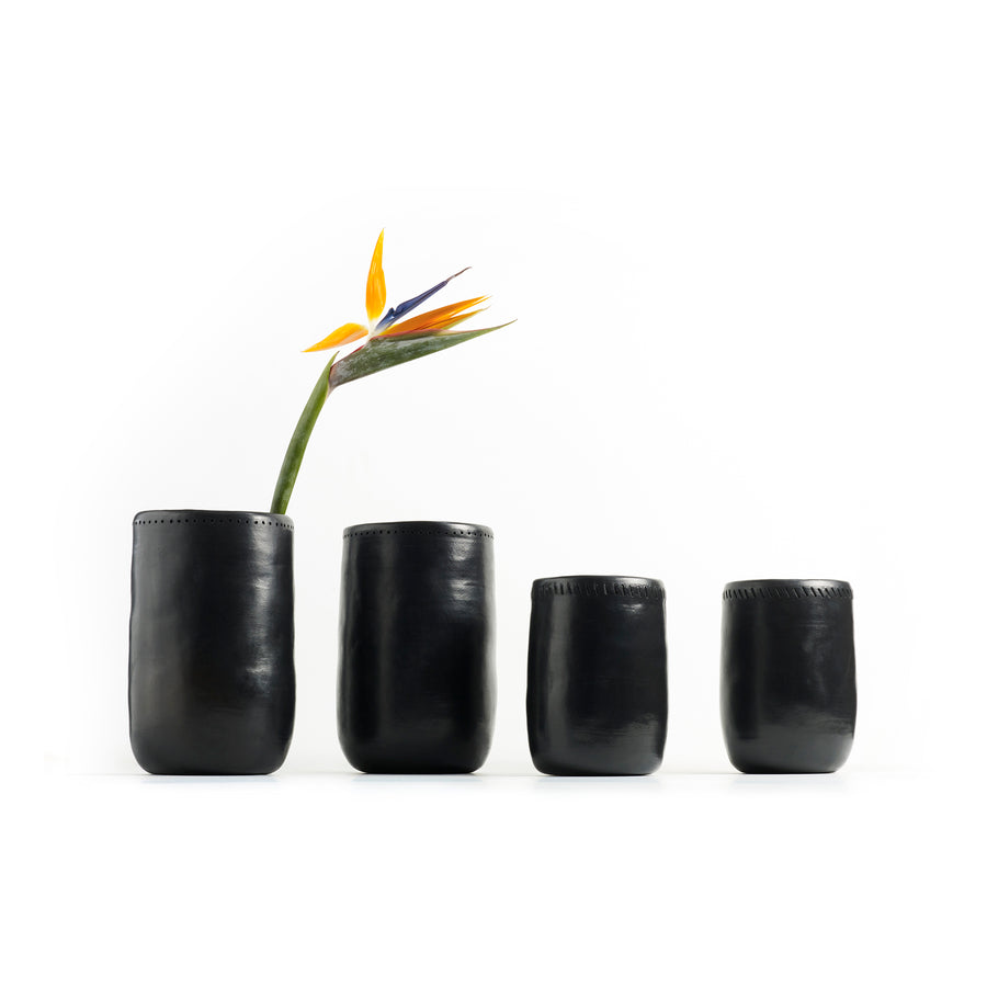 Four Ames Barro Vase 1s in black leather, inspired by handmade pottery, line up against a white background. The tallest vase displays Colombian craftsmanship with a single bird of paradise flower, its vibrant orange and blue petals providing a striking contrast.