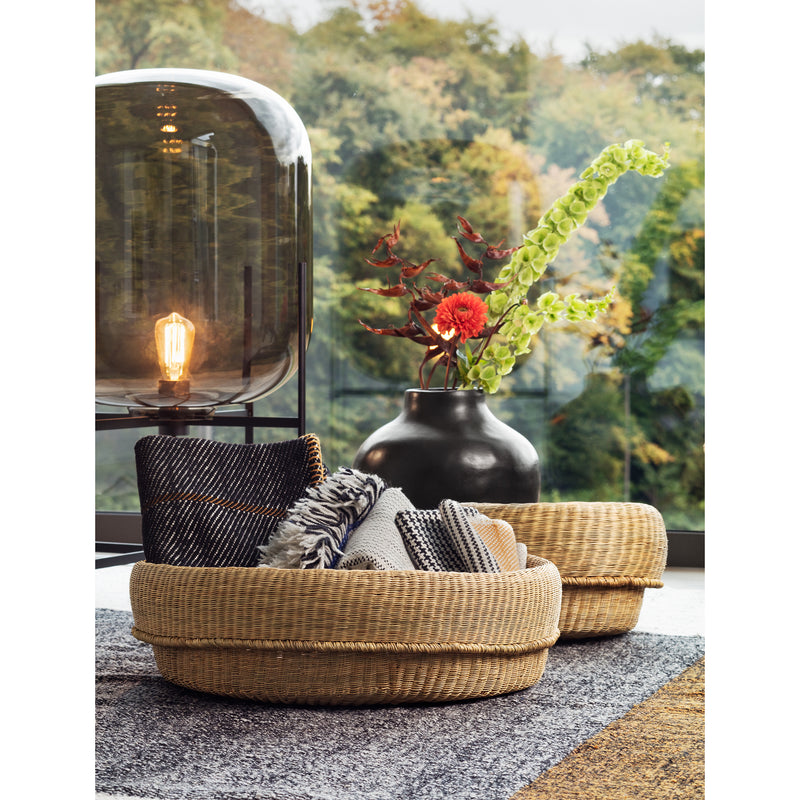 A cozy living room scene features two Ames Fibra Basket Smalls, handcrafted in Colombia, filled with blankets. A black vase with vibrant flowers sits on the table beside a glowing large glass lamp with an exposed bulb, while lush greenery is seen through the window.