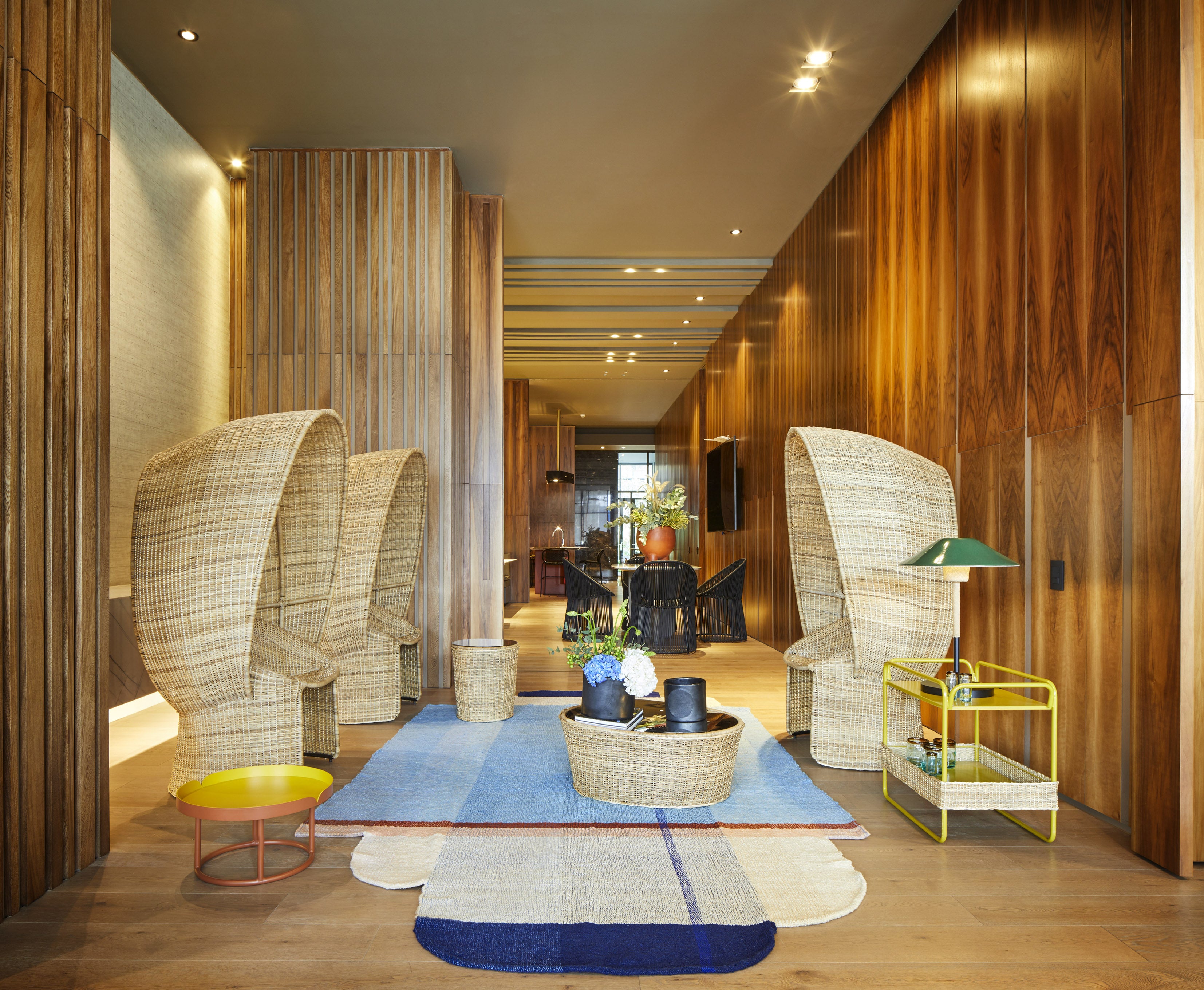 A modern lounge area includes Sebastian Herkners wicker pod chairs, a colorful geometric rug, and small tables with decorative items. Wood-paneled walls create warmth while Ames Cartagenas Natural Sidetable with a green lamp adds a pop of color.