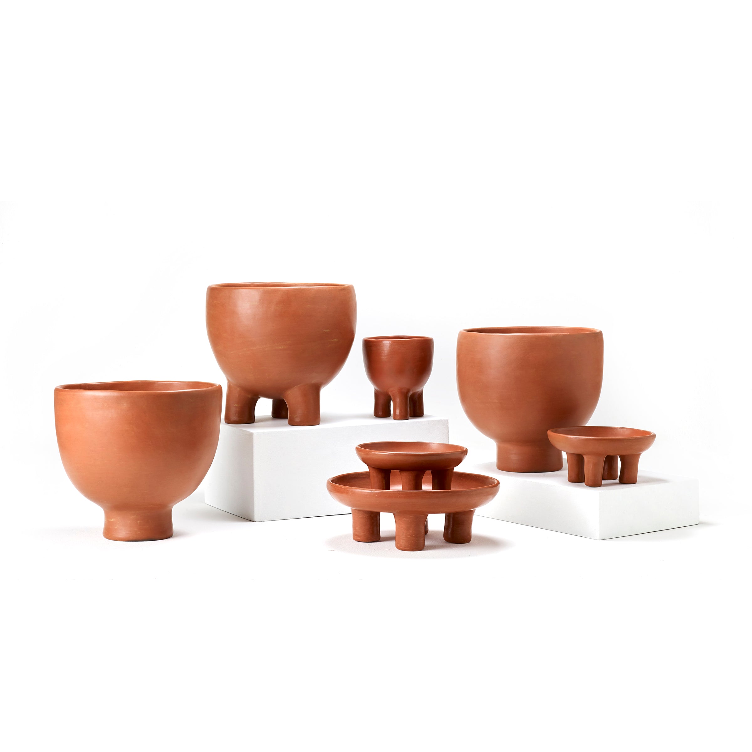 The Ames Barro Tray 1 by Ames features a rounded terracotta form with a tripod base, beautifully displayed on white rectangular blocks to highlight its traditional pottery design against a plain white background.