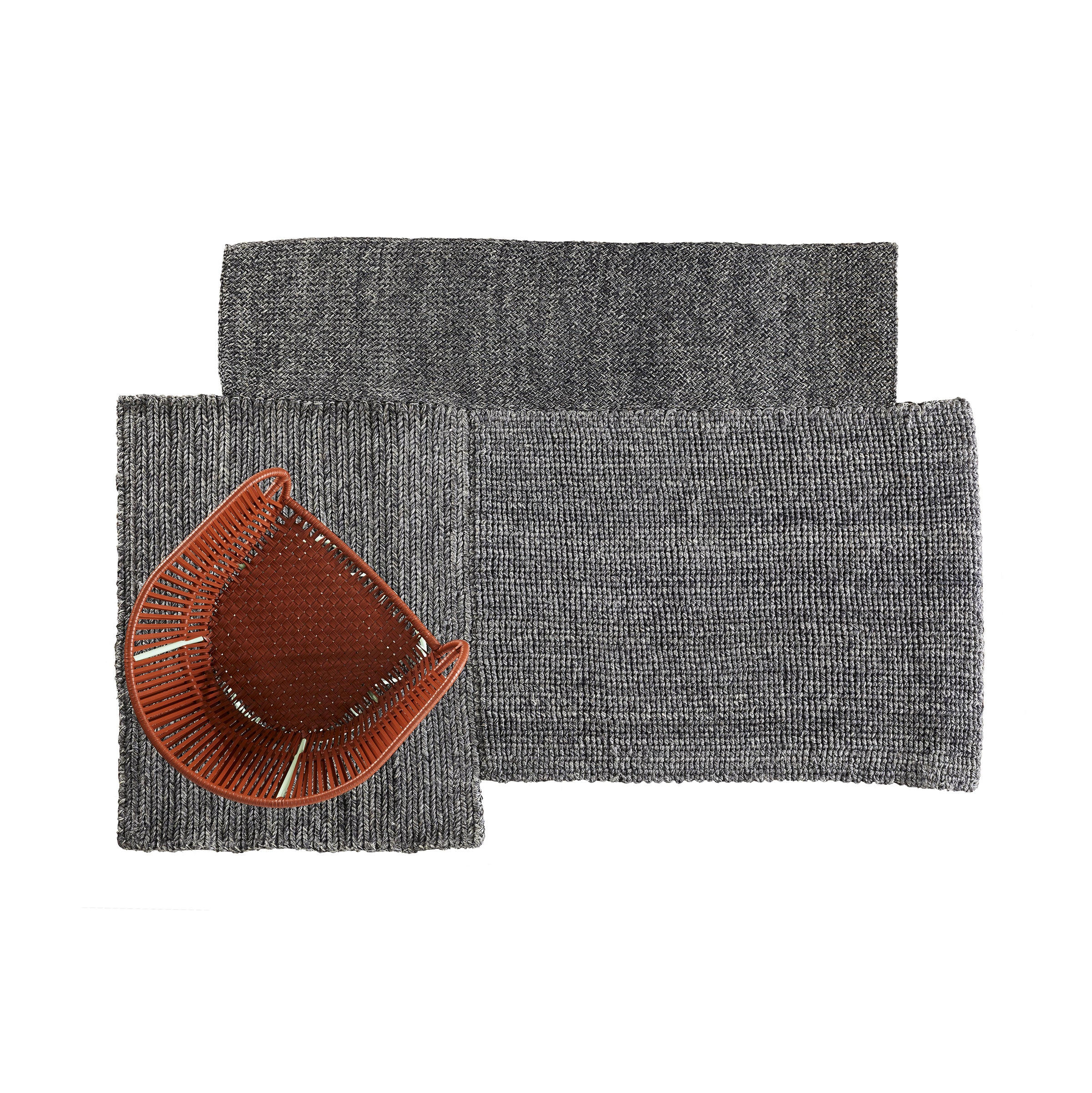 The Ames Cabuya Rug, a woven gray rectangular piece made from flame-retardant fique fiber, elegantly overlaps with a smaller rug. A brown curved vase with vertical ribbing sits on the overlap, contrasting the neutral tones and highlighting Sebastian Herkners design flair.