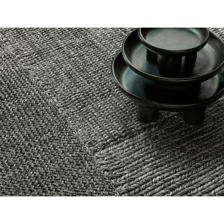 A close-up of the textured gray Cabuya Rug by Ames, featuring various weaving patterns, alongside a set of smooth black ceramic bowls.